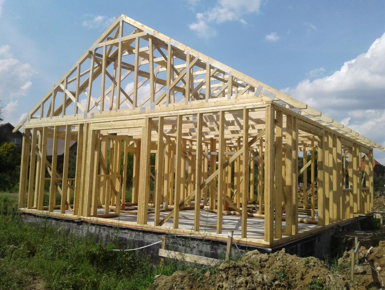 Wznoszenie konstrukcji domu z drewnianych belek, widoczne metalowe łączniki, fundament z betonu, w tle zieleń i błękitne niebo z chmurami.