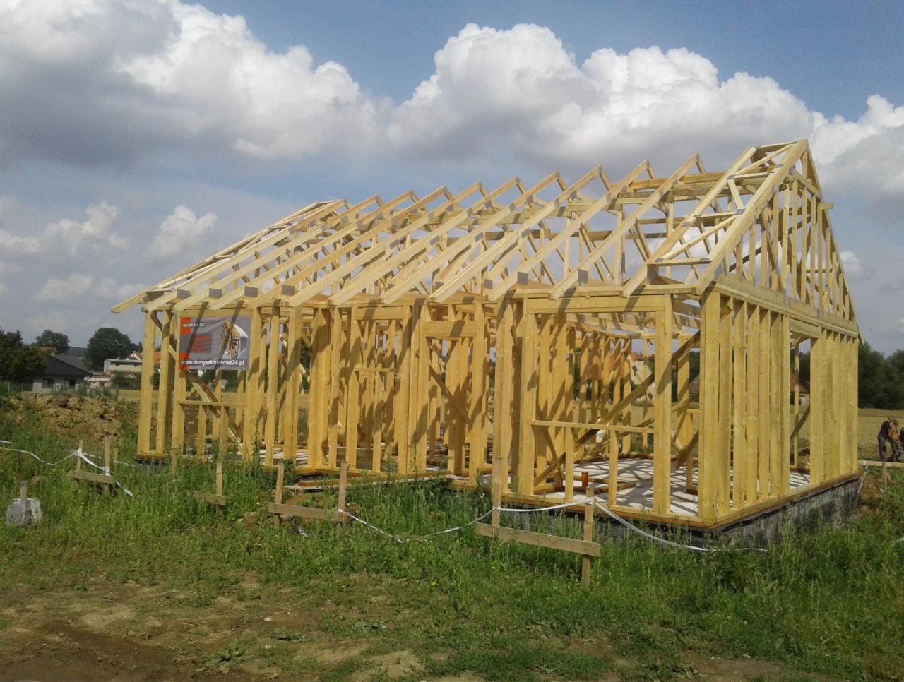 Surowa konstrukcja drewniana domu w trakcie budowy, widoczne fundamenty i szkielet ścian oraz dachu, na pierwszym planie trawa, w tle błękitne niebo z chmurami.