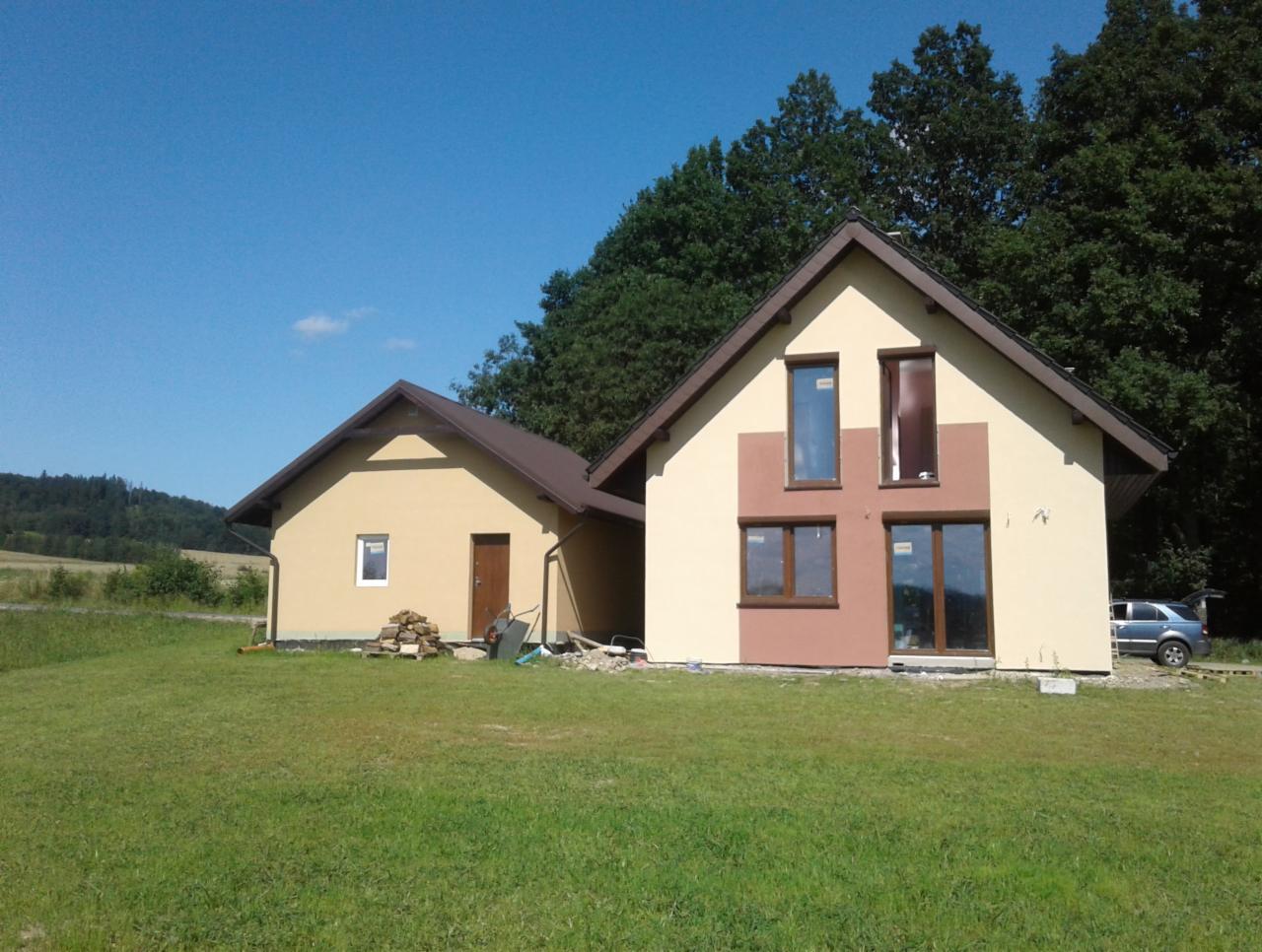 Dwa budynki: parterowy z brązowym dachem i stertą drewna obok wejścia oraz piętrowy z elewacją w odcieniach beżu i brązu, z otwartym bagażnikiem samochodu w tle.