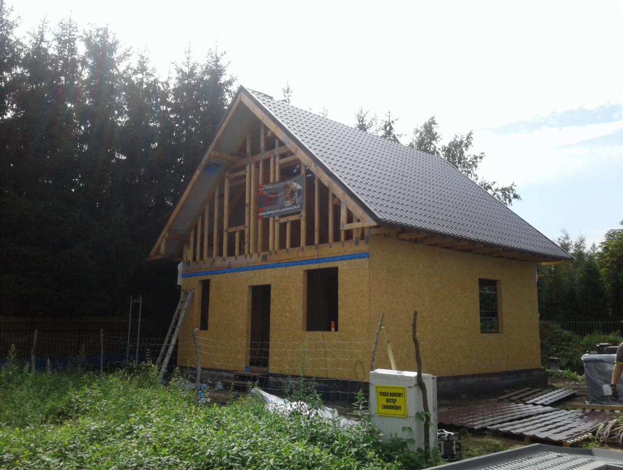 Wznoszony dwukondygnacyjny budynek z drewnianą konstrukcją szkieletową, częściowo obłożony płytami OSB i pokryty ciemną dachówką, widoczny na tle lasu i ogrodzenia.