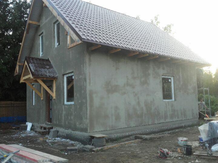 Niedokończony dom murowany z szarą elewacją w trakcie tynkowania, widoczne drewniane elementy konstrukcyjne dachu, okna z białymi ramami, teren budowy z narzędziami i materiałami.