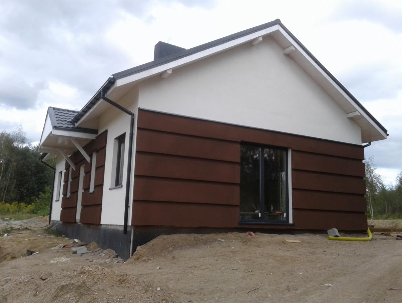 Niedokończony dom z brązową elewacją z poziomych paneli i białym wykończeniem, widoczne fundamenty, rury drenażowe i sterta ziemi przed budynkiem.
