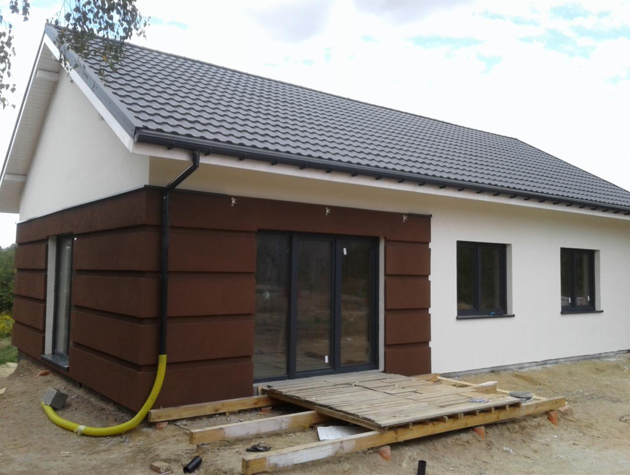 Nowo wybudowany dom z brązową elewacją w pasy i białymi ścianami, z widoczną żółtą rurą na zewnątrz i prowizorycznym drewnianym tarasem.