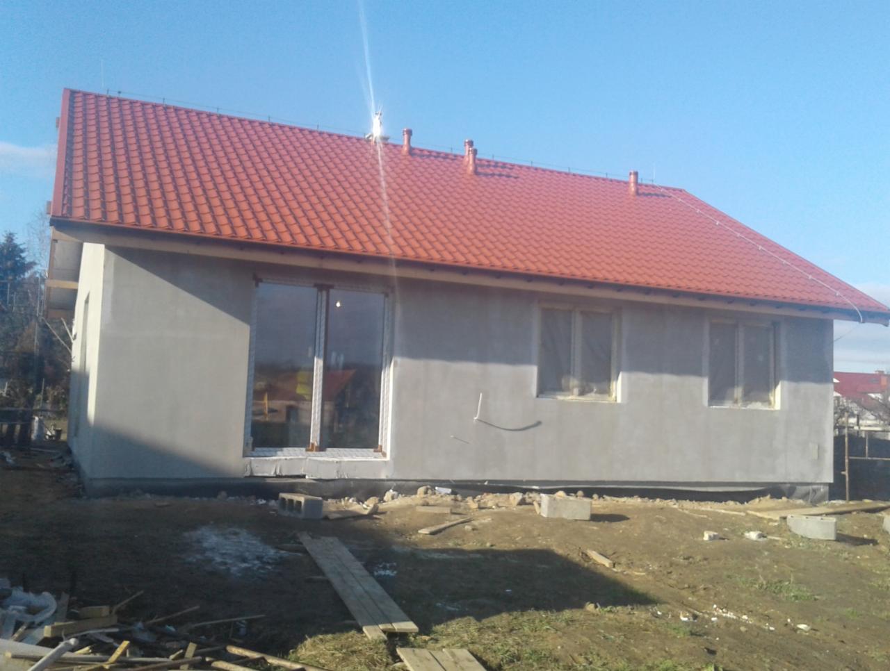 Niedokończony budynek jednorodzinny z czerwoną dachówką, szarymi ścianami zewnętrznymi przed tynkowaniem, dużym oknem tarasowym i widocznymi elementami budowlanymi na trawniku.
