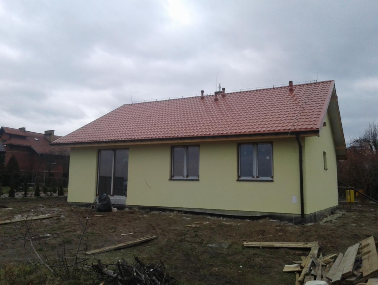 Parterowy dom jednorodzinny z czerwoną dachówką, świeżo otynkowany na żółto, z dużymi oknami, częściowo zakryty folią, na działce w trakcie prac wykończeniowych, pochmurne niebo.