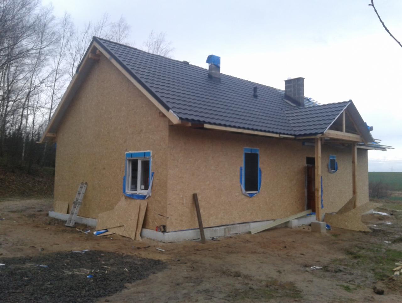 Surowy stan budynku w trakcie budowy z płyt OSB, czarna dachówka, widoczne zabezpieczenia okien folią, teren budowy z gruzem i piachem.