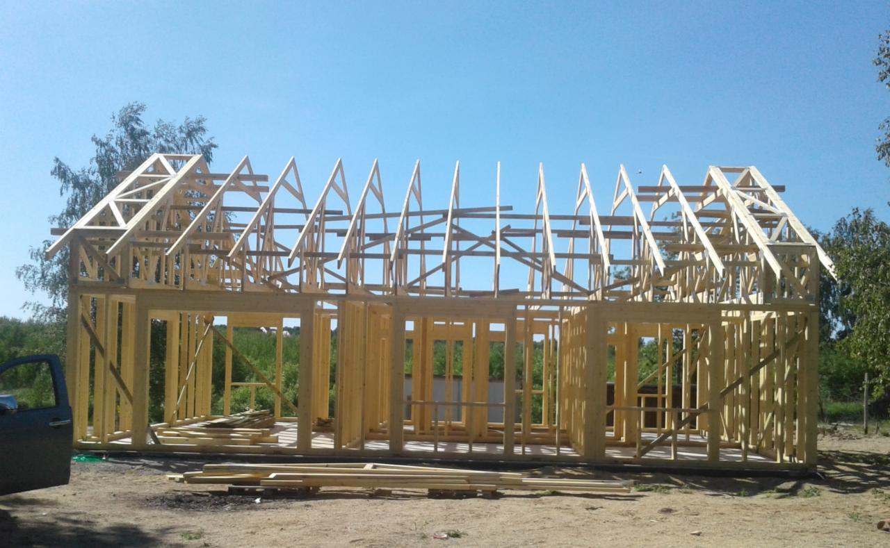 Konstrukcja szkieletowa domu w trakcie budowy, widoczne drewniane elementy ścian i dachu, teren budowy z zielenią w tle, fragment samochodu po lewej stronie.
