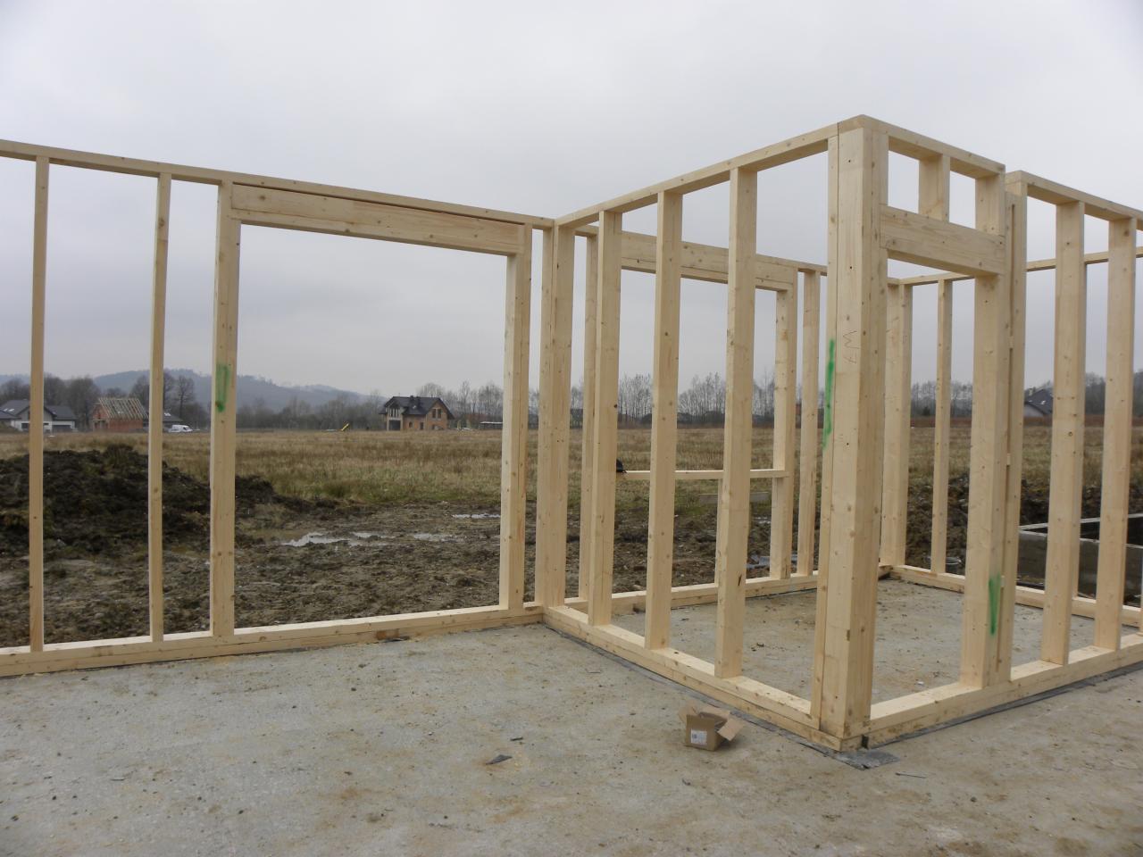 Surowa konstrukcja ścian z drewna na fundamencie, widok na krajobraz z domami w tle, pochmurny dzień.