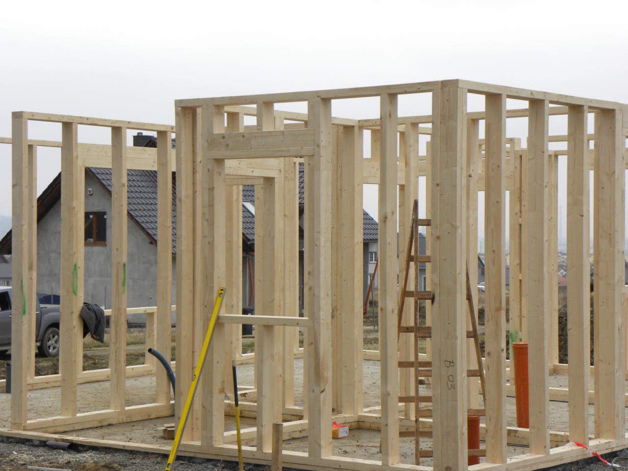 Wznoszenie konstrukcji domu z drewnianych belek, widoczne narzędzia budowlane i drabina, w tle zabudowa jednorodzinna.