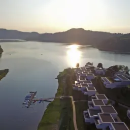 Widok z lotu ptaka na kompleks apartament&oacute;w z płaskimi dachami nad jeziorem, w tle zalesione wzg&oacute;rza i zachodzące słońce odbijające się w wodzie.