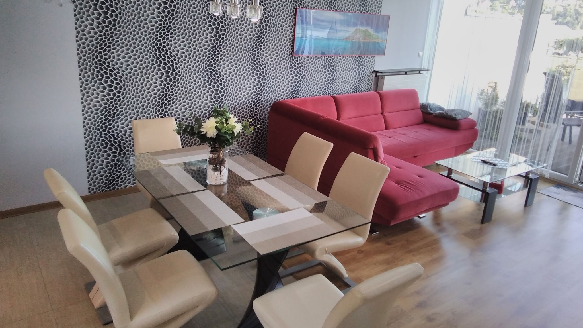 Jasny salon z czerwonym narożnikiem, szklanym stolikiem kawowym i stołem jadalnym z kwiatami na tle wzorzystej tapety.