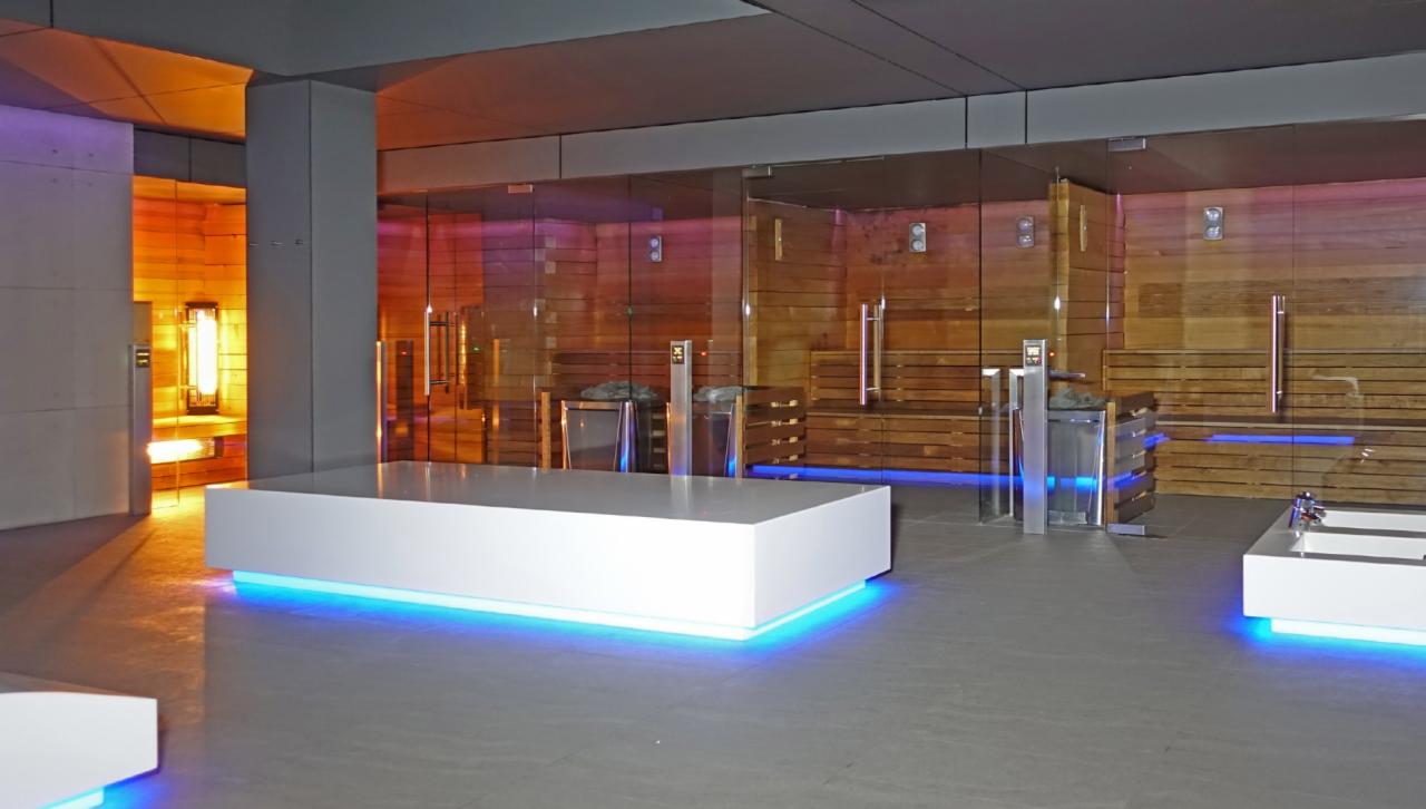 Nowoczesne wnętrze strefy SPA z saunami za szklanymi ścianami, podświetlanymi białymi leżankami i elementami dekoracyjnymi z niebieskim oświetleniem LED.