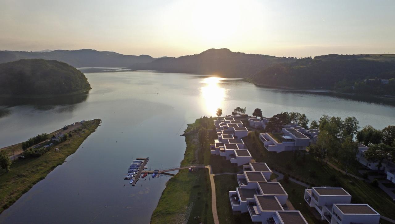 Widok z lotu ptaka na kompleks apartamentów z płaskimi dachami nad jeziorem, w tle zalesione wzgórza i zachodzące słońce odbijające się w wodzie.