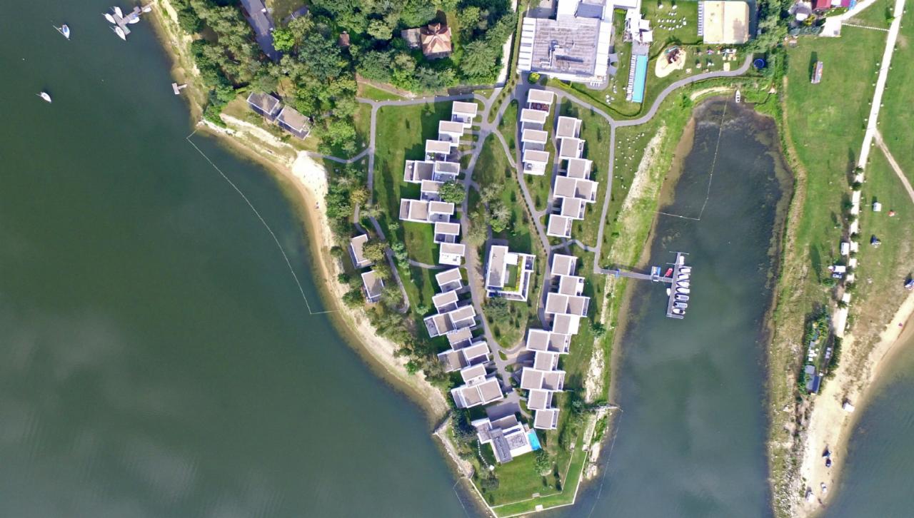 Widok z lotu ptaka na kompleks apartamentów wypoczynkowych z białymi, nowoczesnymi budynkami położonymi nad jeziorem, otoczonych zielenią, z prywatną plażą i przystanią dla łodzi.