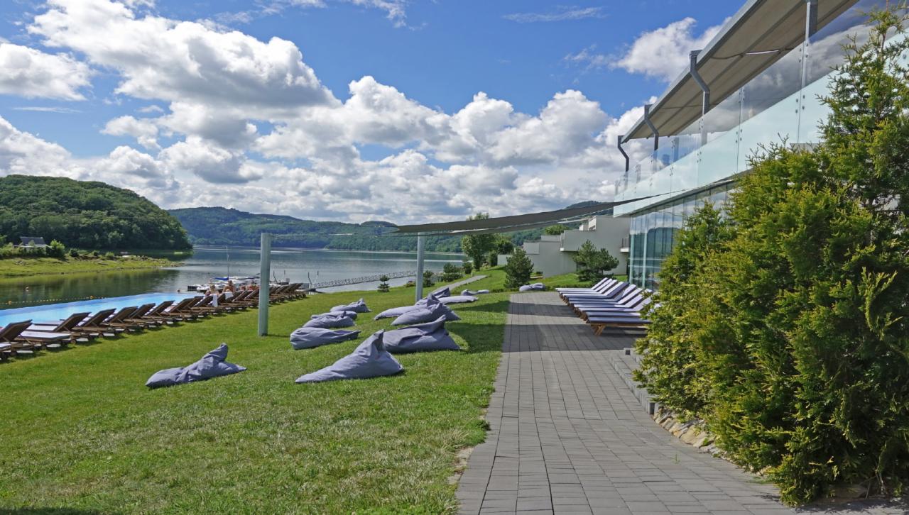 Widok na jezioro z terenu Lemon Resort SPA: basen, leżaki, pufy na trawie, nowoczesny budynek z przeszkloną fasadą.