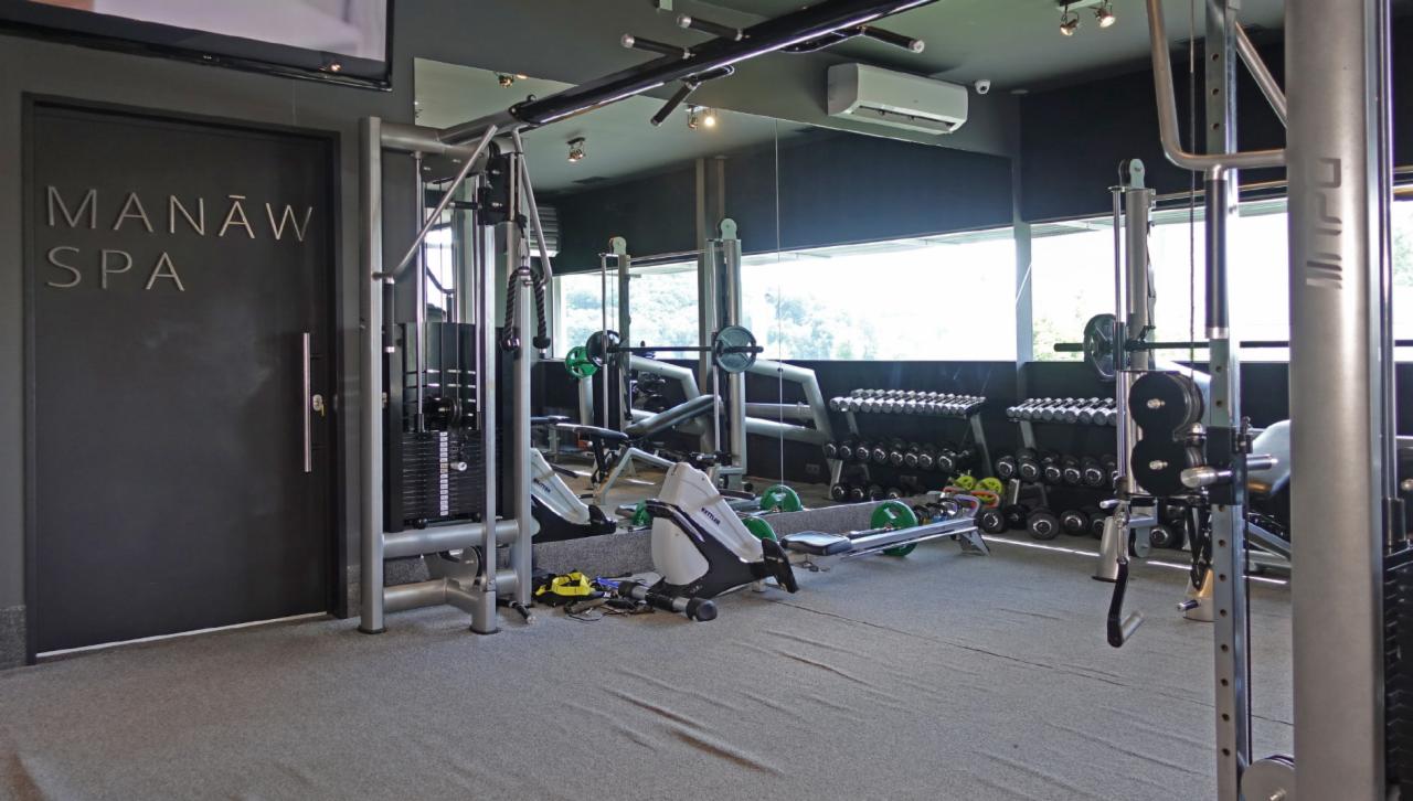 Nowoczesna siłownia w SPA z różnorodnym sprzętem do ćwiczeń siłowych i cardio, widok na okno i logo Manaw SPA na drzwiach.