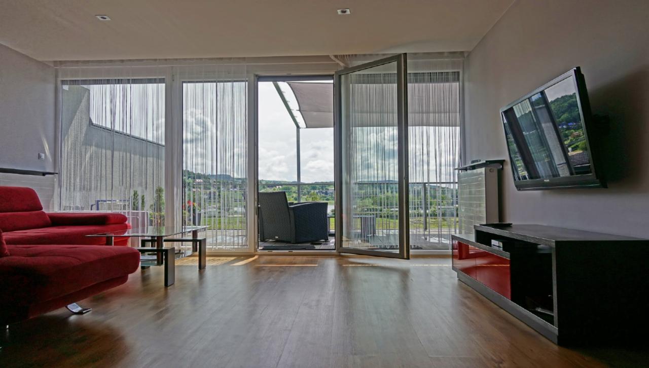 Jasne wnętrze apartamentu z dużym oknem balkonowym, widok na zielone wzgórza, czerwona sofa i szklany stolik kawowy, telewizor na ścianie.