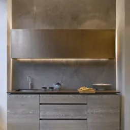 Minimalistyczna kuchnia z drewnianymi szafkami, szarym tłem i subtelnym oświetleniem LED pod metalową półką, na blacie czajnik i naczynia.