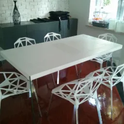 Biały stół z krzesłami o geometrycznym wzorze na tle ściany z białej cegły i ciemnej szafki, widok na okno z klatką dla ptaków.