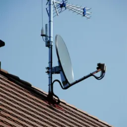 Antena satelitarna i antena telewizyjna zamontowane na metalowym maszcie na dachu pokrytym czerwoną dachówką, widok z dołu na tle błękitnego nieba.