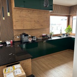 Przedsiębiorstwo Handlowo Usługowe Drewmir Marcin Miraszewski - Nowoczesna kuchnia z drewnianymi frontami szafek górnych i ciemnozielonymi szafkami dolnymi, czarnym blatem, drewnianą okładziną ścienną i złotymi akcentami, widoczne kartony na podłodze.