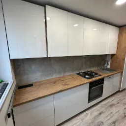 Nowoczesna kuchnia z białymi, błyszczącymi szafkami górnymi i dolnymi, drewnianym blatem i szarą płytką na ścianie, widoczne narzędzia montażowe przy oknie.