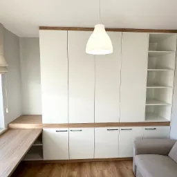 Nowoczesna szafa wnękowa z białymi frontami i drewnianymi akcentami, połączona z biurkiem przy oknie, oświetlona wiszącą lampą, uzupełniona o jasną sofę.