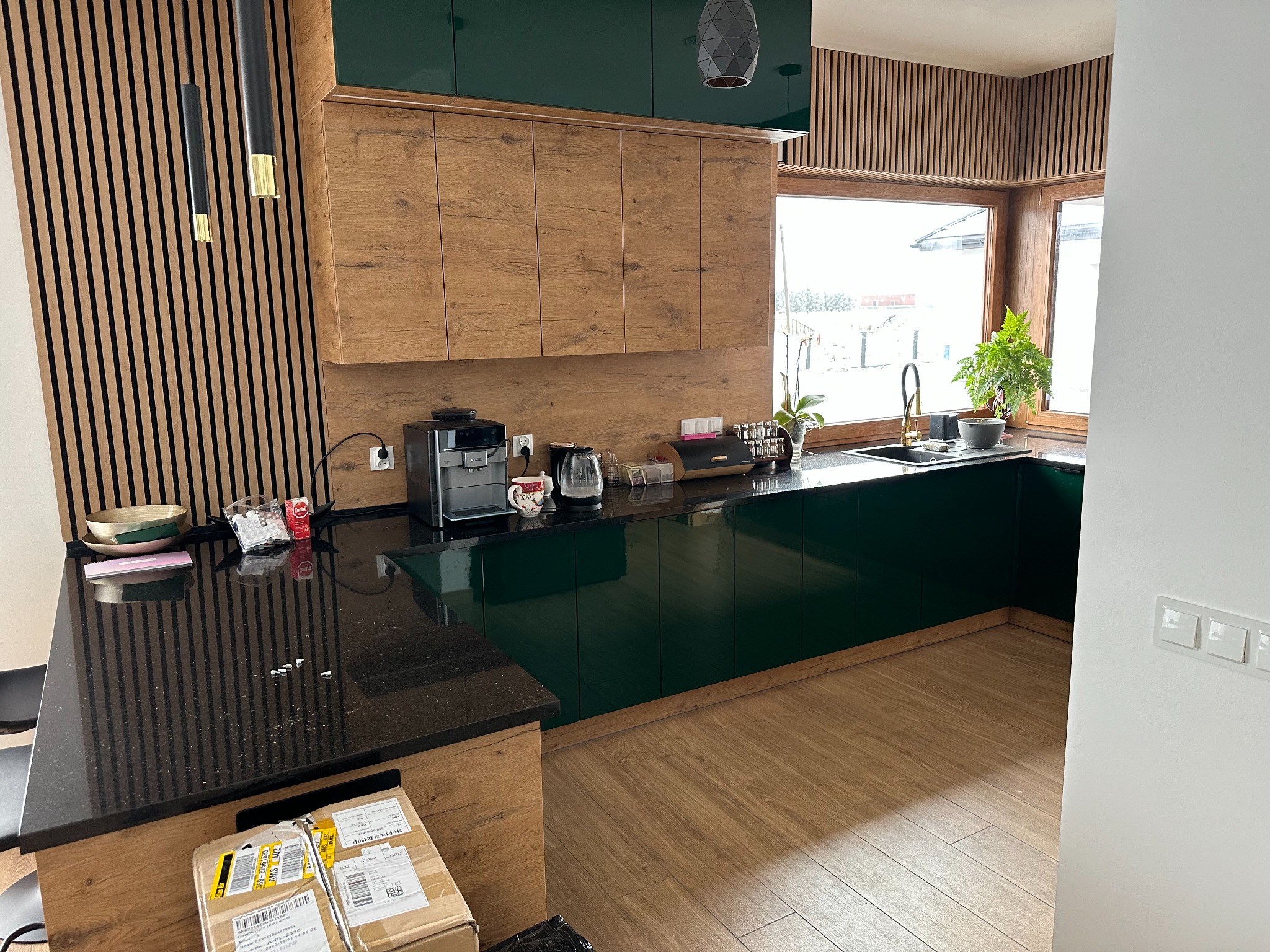 Nowoczesna kuchnia z drewnianymi frontami szafek górnych i ciemnozielonymi szafkami dolnymi, czarnym blatem, drewnianą okładziną ścienną i złotymi akcentami, widoczne kartony na podłodze.