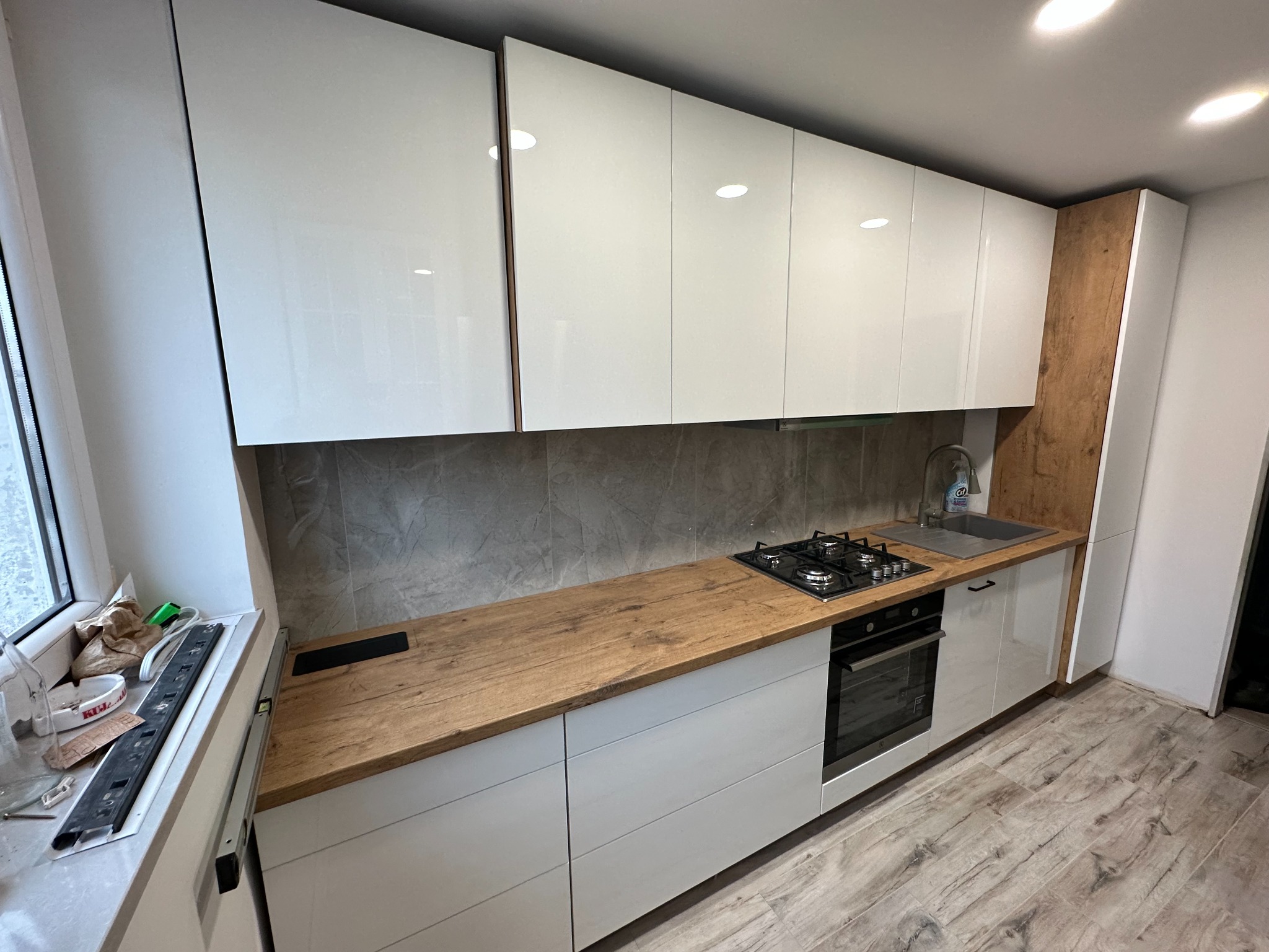 Nowoczesna kuchnia z białymi, błyszczącymi szafkami górnymi i dolnymi, drewnianym blatem i szarą płytką na ścianie, widoczne narzędzia montażowe przy oknie.