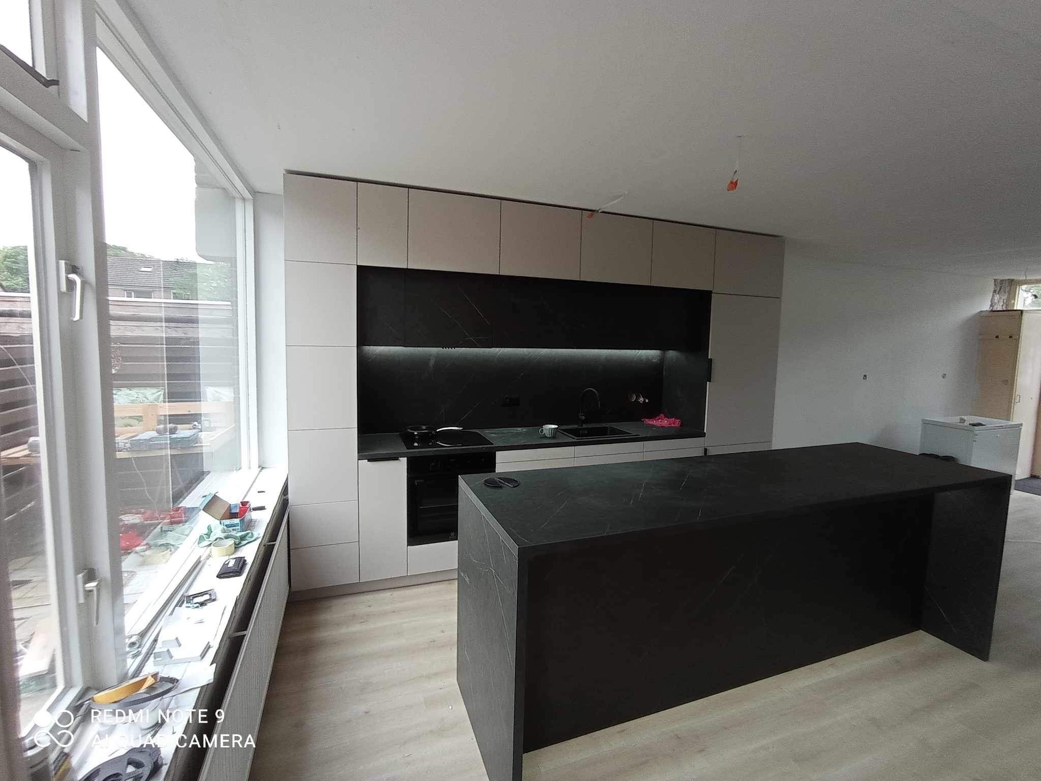 Nowoczesna kuchnia z wyspą, jasne szafki górne i czarny panel ścienny z marmurkowym wzorem, widok z okna z narzędziami i materiałami budowlanymi na parapecie, w trakcie wykańczania.