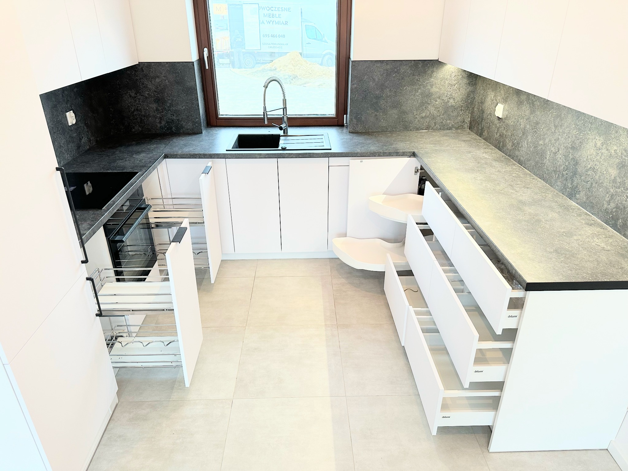 Nowoczesna, biała kuchnia z szarym blatem i otwartymi szufladami cargo oraz narożną szafką z półkami obrotowymi, widok przez okno na plac budowy.