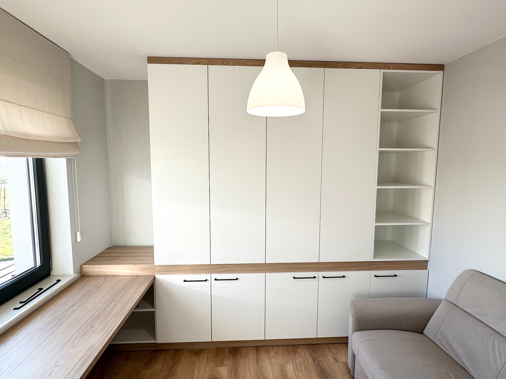 Nowoczesna szafa wnękowa z białymi frontami i drewnianymi akcentami, połączona z biurkiem przy oknie, oświetlona wiszącą lampą, uzupełniona o jasną sofę.