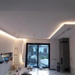 Wnętrze pokoju w trakcie prac wykończeniowych z podwieszanym sufitem z oświetleniem LED, widoczna drabina malarska, kartony i narzędzia na podłodze. Ściana z cegły pomalowana na biało i czarny...