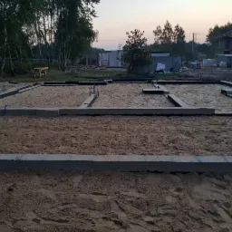 Fundamenty pod dom jednorodzinny w trakcie budowy, widoczne zbrojenia i szalunki, w tle drzewa i inne budynki w stanie surowym.