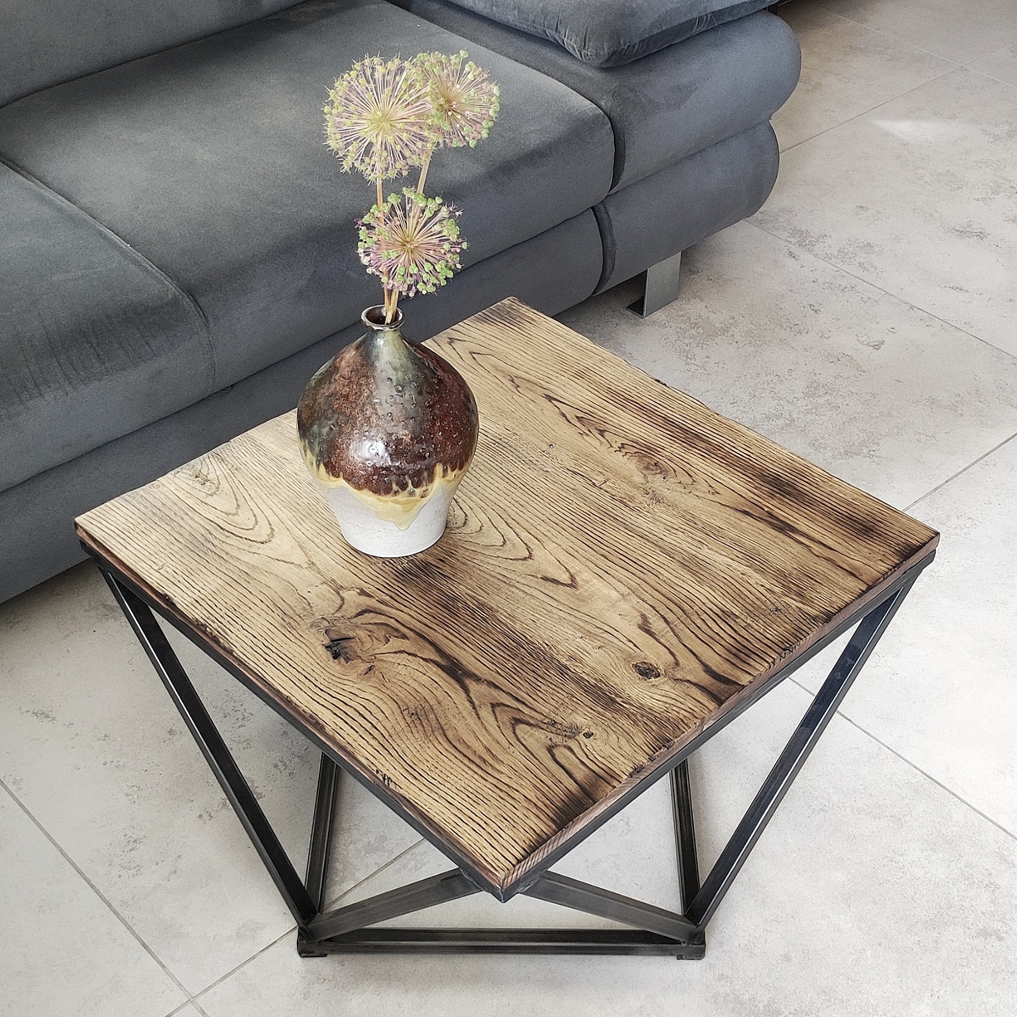 Kwadratowy stolik kawowy z blatem z postarzanego drewna i metalową podstawą w kształcie rombu, na blacie stoi ceramiczny wazon z kulistymi kwiatami, w tle szara sofa.
