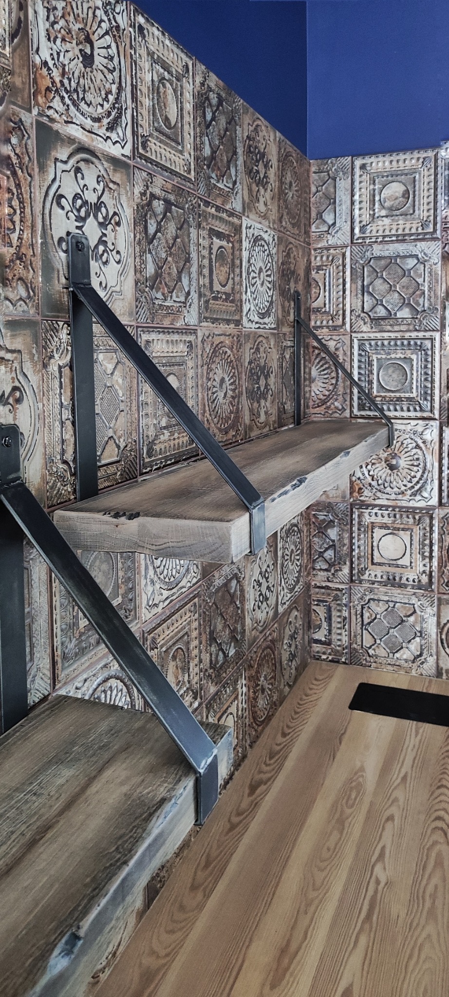 Dwie drewniane półki zamocowane na metalowych wspornikach na tle ściany wyłożonej dekoracyjnymi płytkami w stylu vintage.