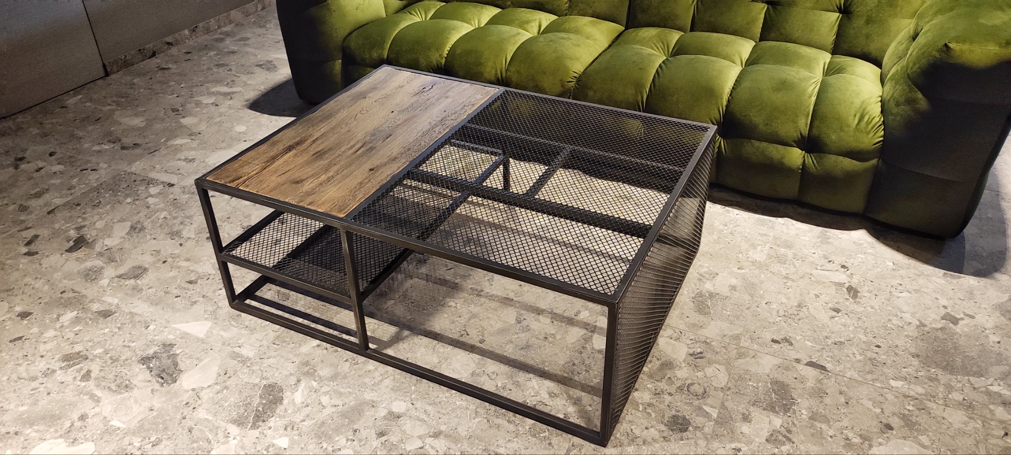 Stolik kawowy z metalową konstrukcją i blatem częściowo z drewna, stojący na marmurowej podłodze, z zieloną sofą w tle.