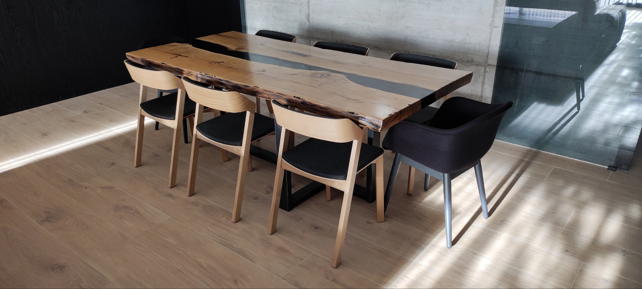 Stół z blatem z naturalnego drewna z żywicą epoksydową w kolorze szarym, ustawiony w nowoczesnym wnętrzu z drewnianą podłogą i krzesłami z jasnego drewna i ciemnymi siedziskami.