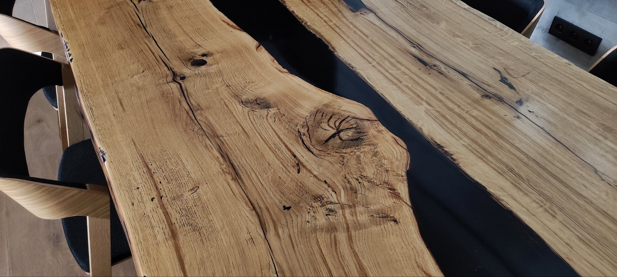 Blat stołu z naturalnego drewna z żywicą epoksydową w kolorze czarnym, widoczne słoje i sęki, nowoczesne krzesła w tle.