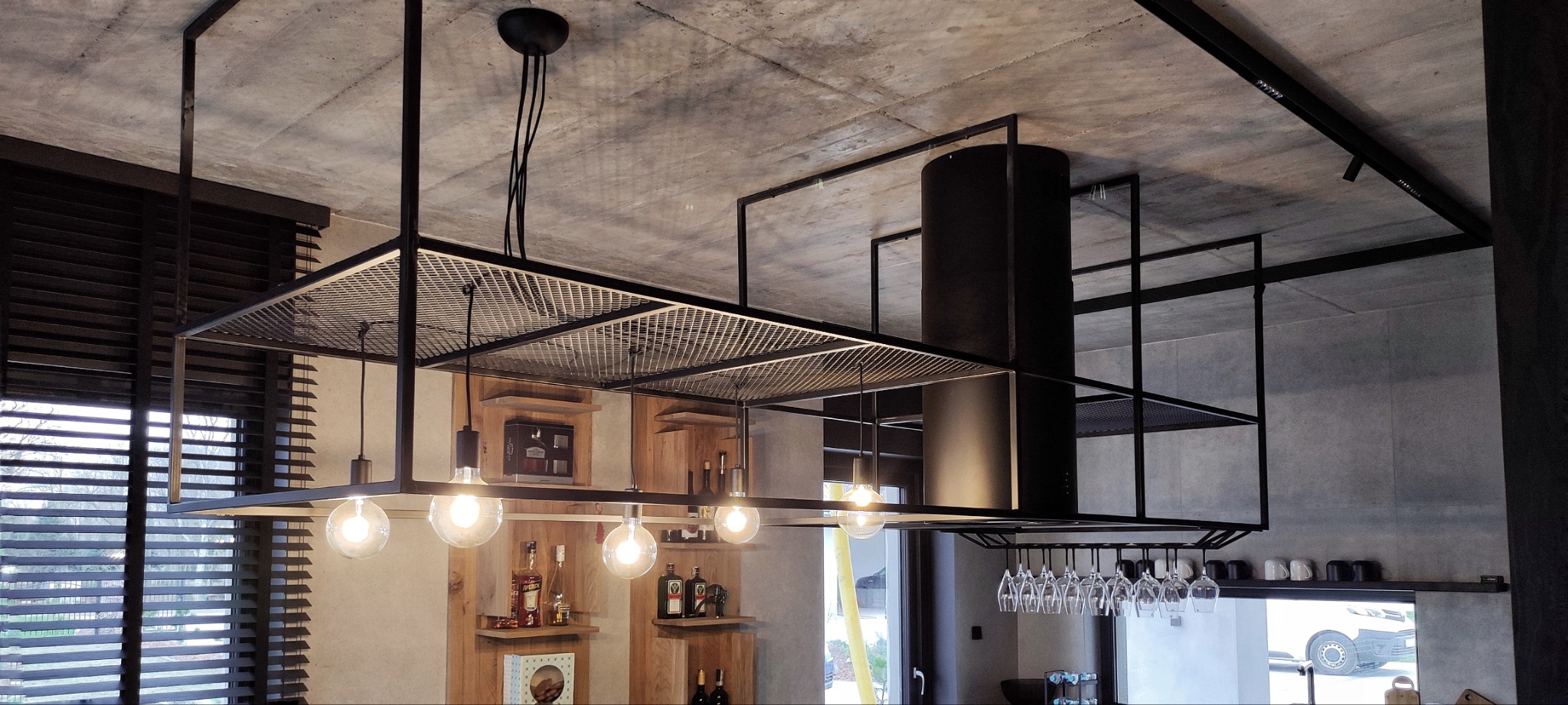 Nowoczesna kuchnia z wyspą o metalowej konstrukcji, na której zawieszone są lampy z żarówkami Edisona oraz okap, widoczne półki z butelkami alkoholi i szklanki do wina.