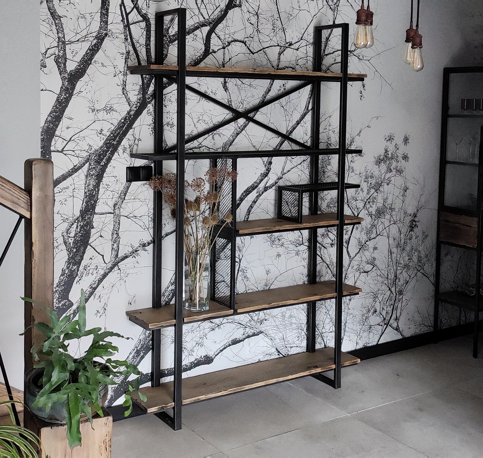 Regał w stylu industrialnym z czarną metalową konstrukcją i drewnianymi półkami, na tle ściany z motywem drzew, z dekoracjami w postaci suszonych kwiatów w wazonie i roślin doniczkowych.