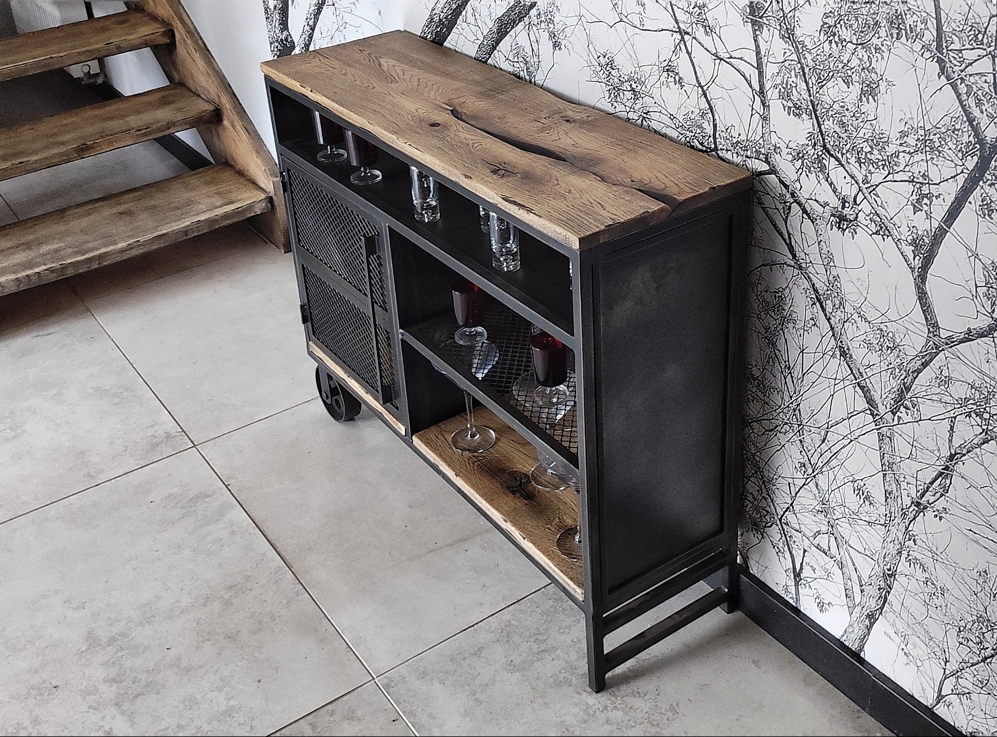Metalowa szafka barowa z drewnianym blatem i ażurowymi drzwiczkami, na kółkach, z kieliszkami w środku, ustawiona na tle ściany z motywem drzewa i drewnianych schodów.