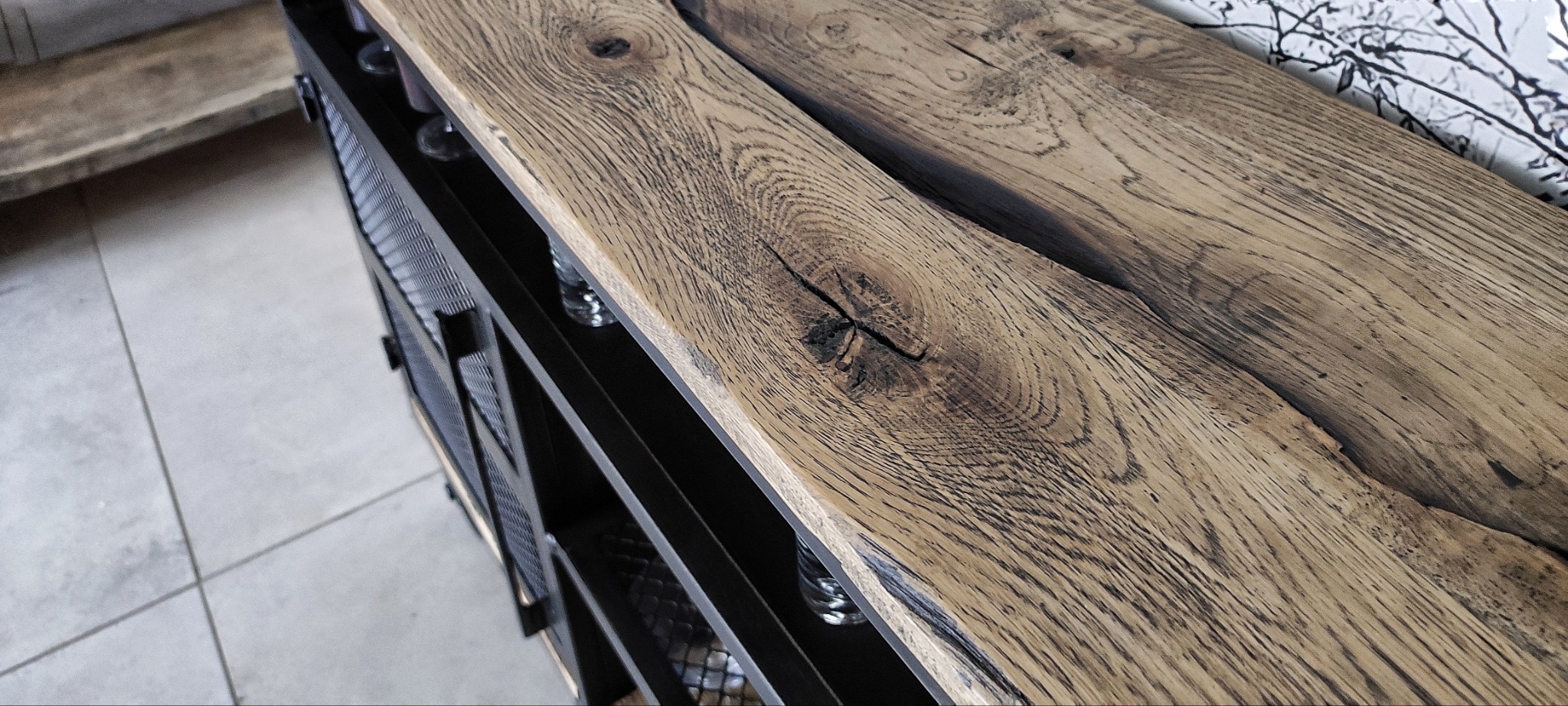 Metalowy regał z blatem z dwóch desek z wyraźnym usłojeniem i sękami, widoczne szklane butelki w przegródkach.