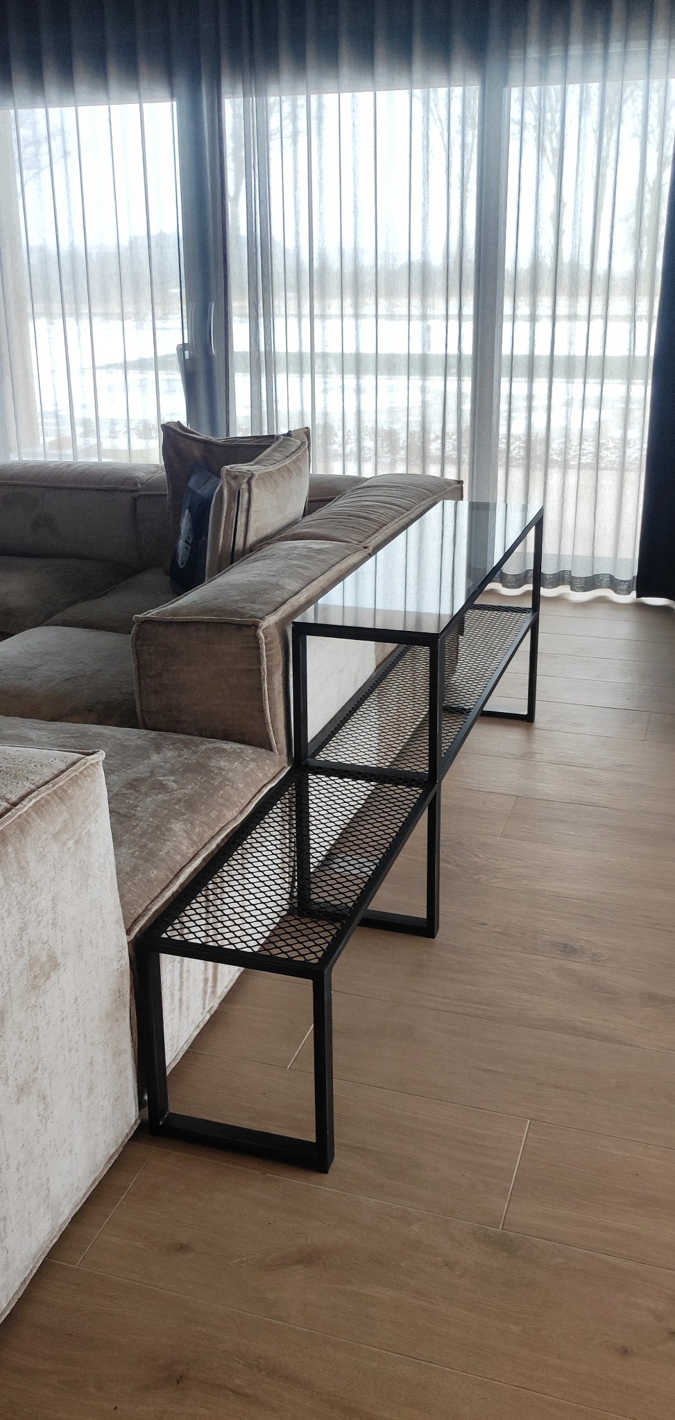 Nowoczesny stolik pomocniczy z czarną metalową konstrukcją i szklanym blatem, przylegający do beżowej, welurowej sofy w jasnym salonie z dużym oknem.