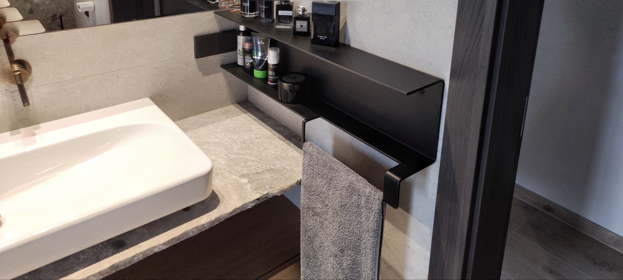 Nowoczesna łazienka w Trzebiatowie z białą umywalką nablatową, kamiennym blatem, ciemnymi półkami na kosmetyki i szarym ręcznikiem.