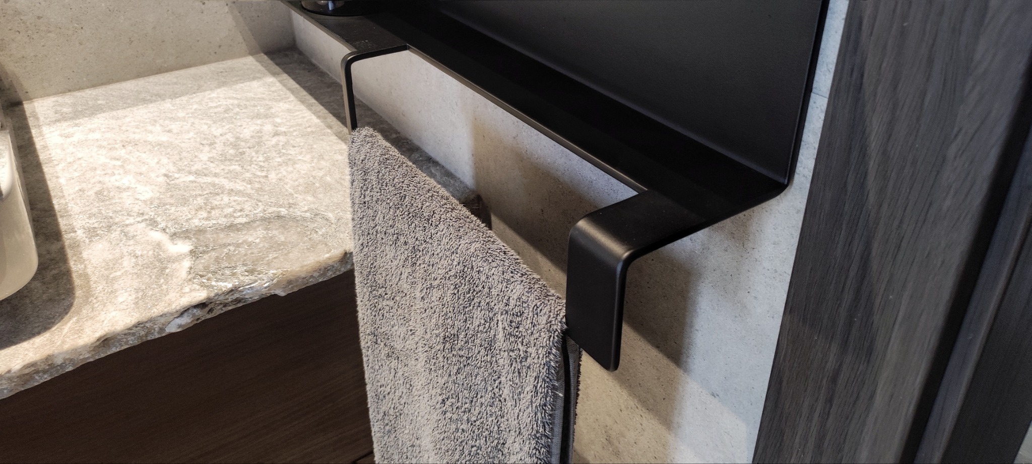 Szary ręcznik wiszący na czarnym, metalowym wieszaku w łazience obok blatu z jasnego kamienia.