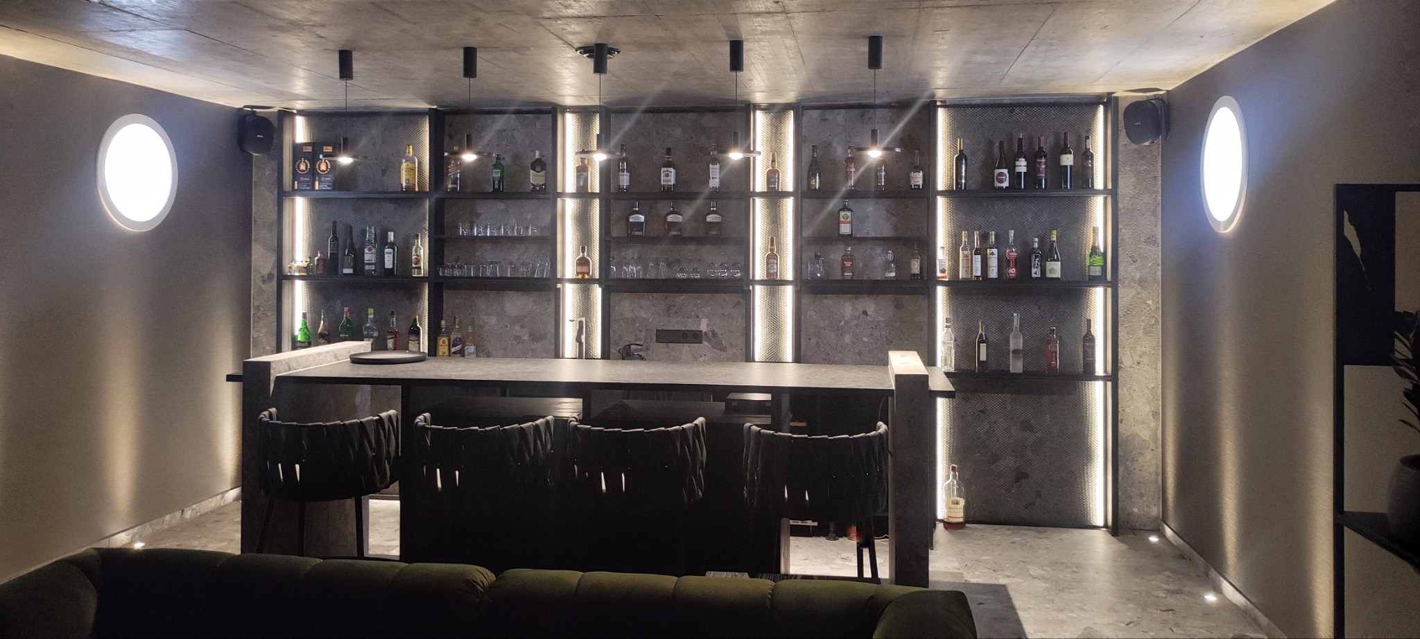 Nowoczesny bar w domu z szarym blatem, krzesłami z plecionki i podświetlanymi półkami wypełnionymi butelkami alkoholi i szklankami. Owalne lampy ścienne po bokach.