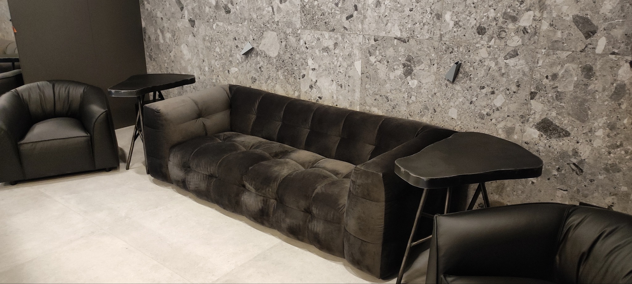 Ciemnoszary, pluszowy zestaw wypoczynkowy: sofa z pikowanymi poduchami i dwa fotele, ustawione na tle ściany wyłożonej płytkami imitującymi kamień.