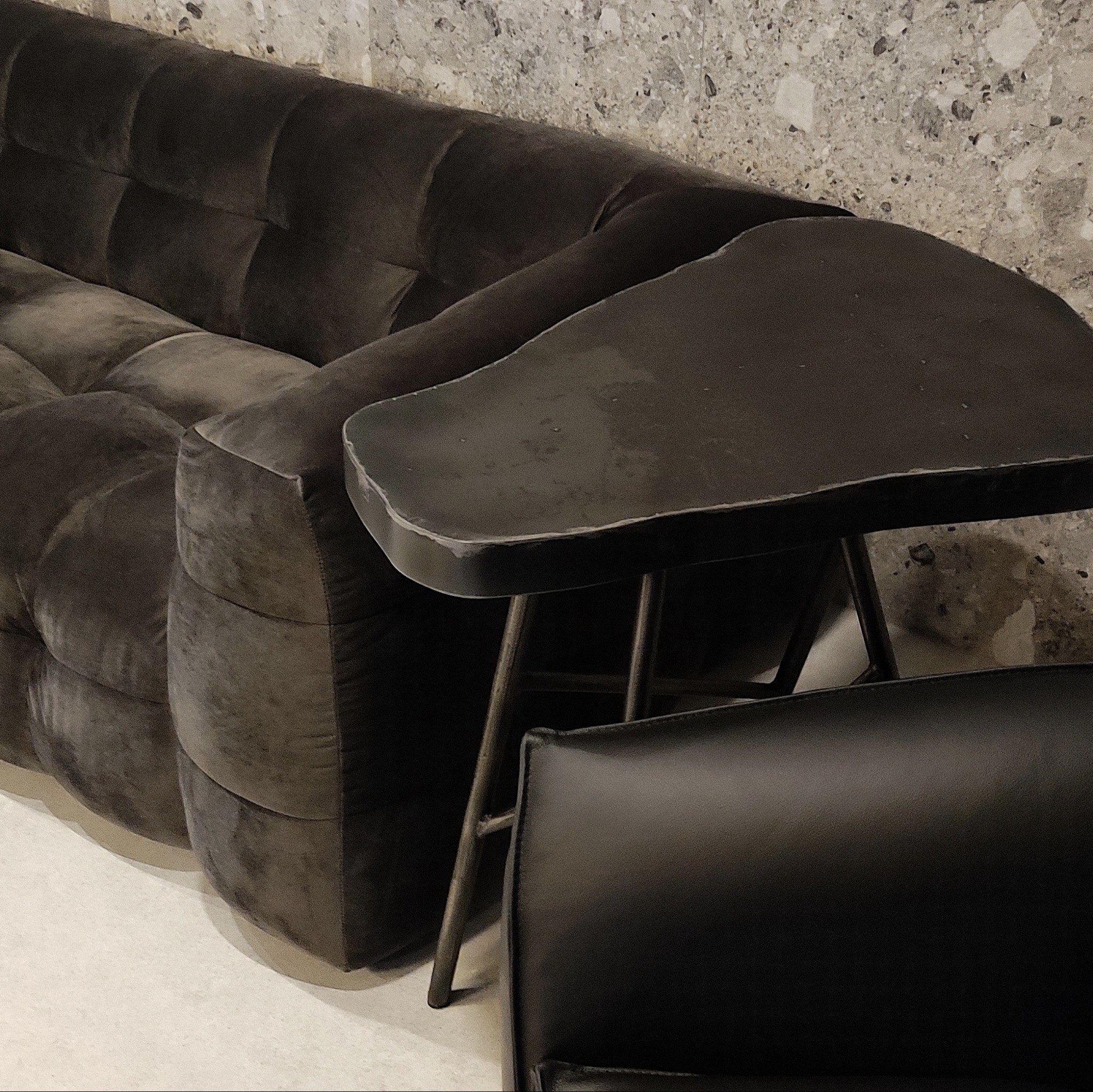 Fragment ciemnobrązowej, pikowanej sofy z podłokietnikiem oraz czarnego skórzanego krzesła i małego stolika na metalowych nogach na tle ściany w plamki.