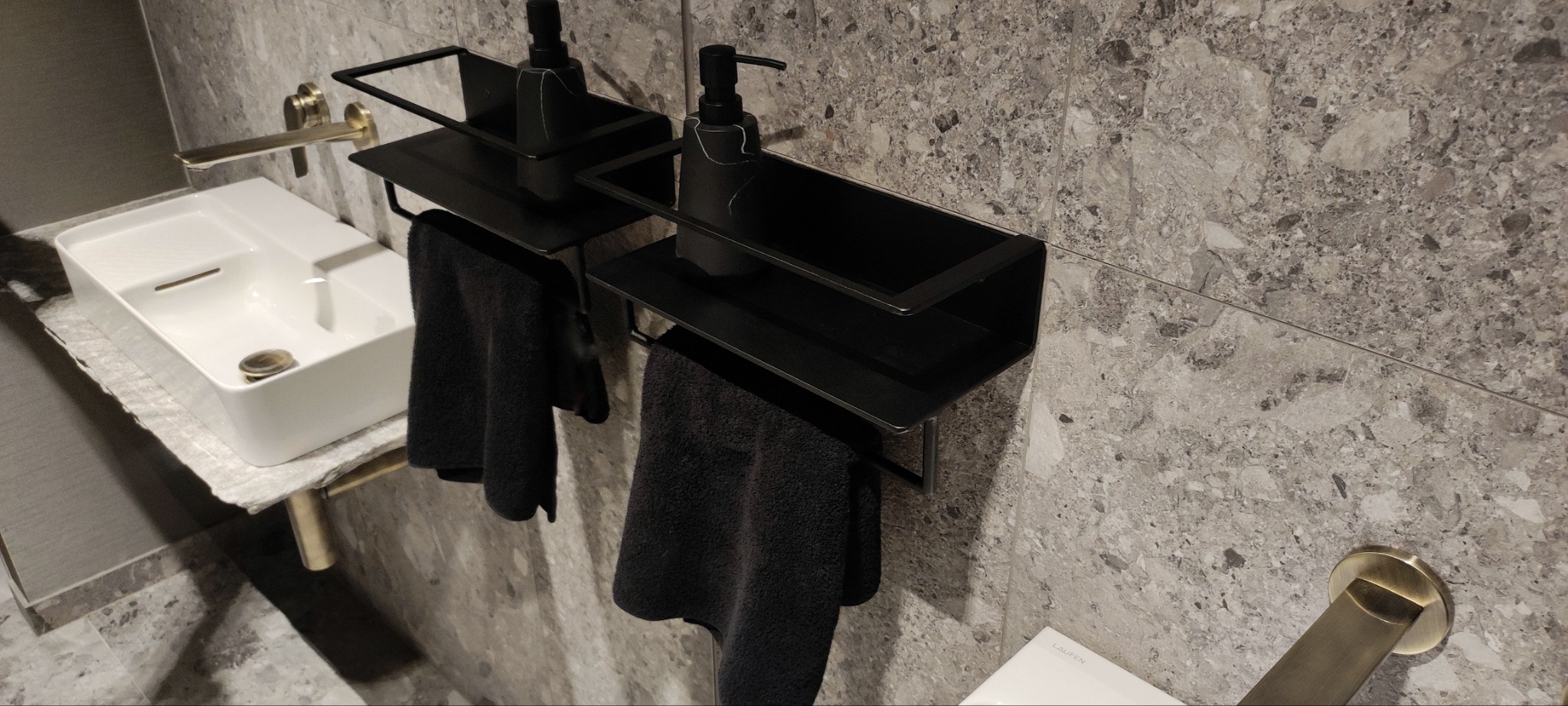 Nowoczesna łazienka z umywalką, złotą armaturą i czarnymi metalowymi półkami na ręczniki, na których stoją dozowniki na mydło.